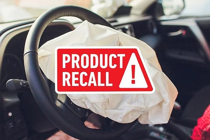 Deployed airbag on a steering wheel behind a red Product Recall warning sign