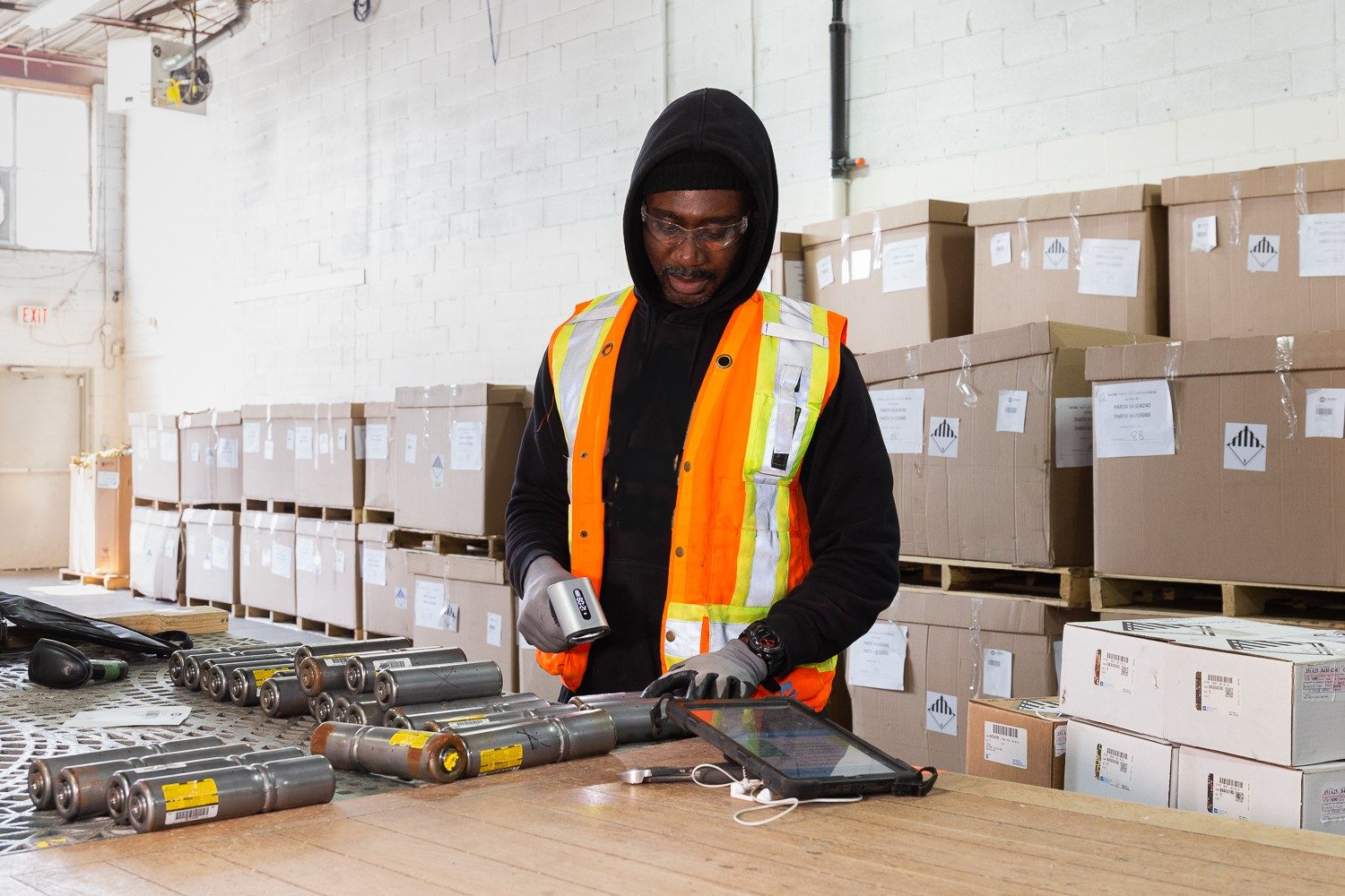 Technician in a hi-vis vest and safety glasses scanning barcodes on airbag inflators, with labelled hazardous-goods boxes behind