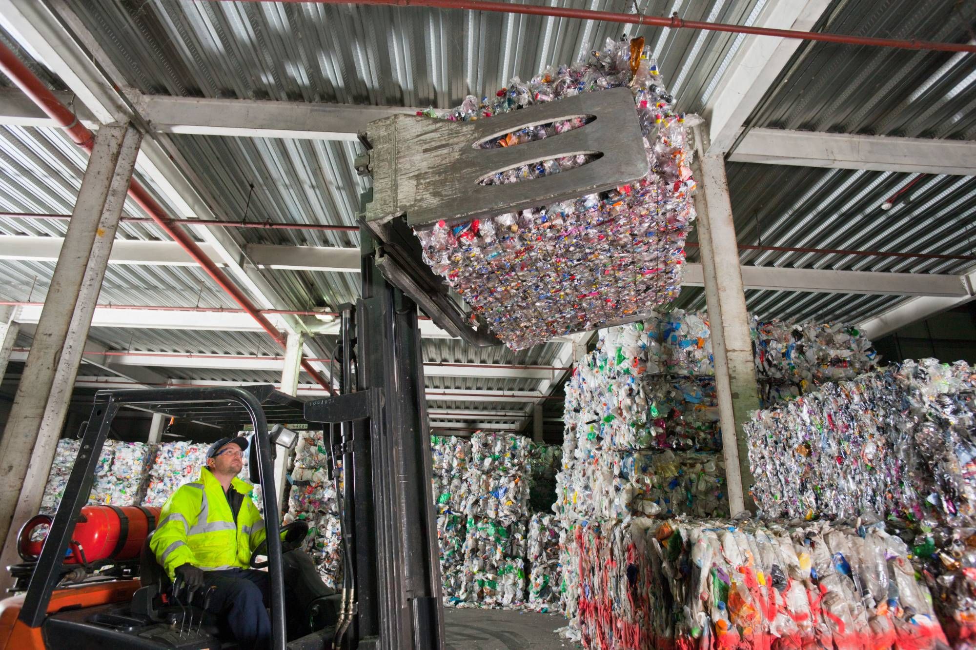Forklift operator in hi-vis lifting a bale of post-industrial plastics in a warehouse stacked with baled material