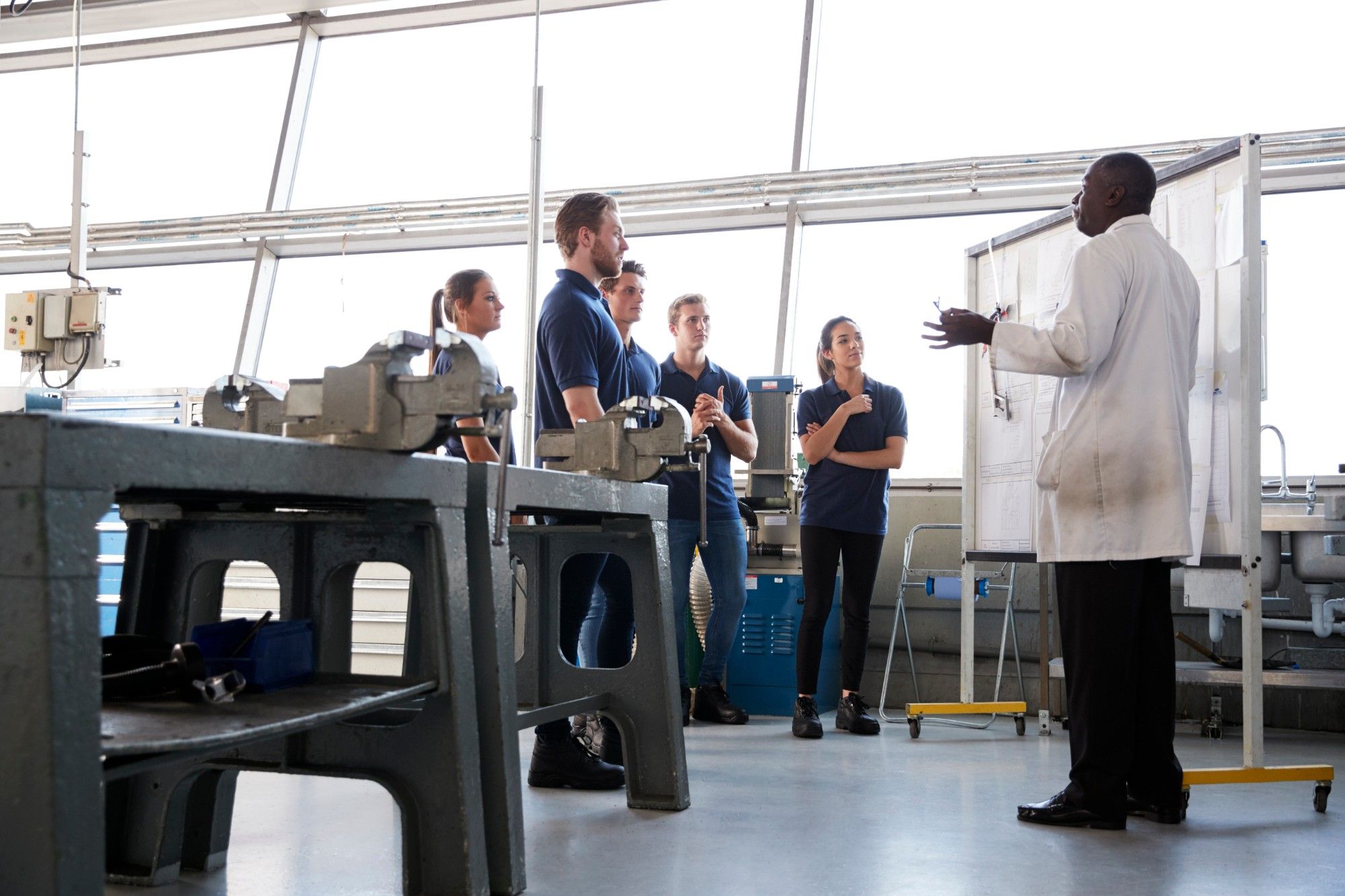 Team members in navy polos listening to an instructor in a lab coat at a whiteboard in a bright workshop