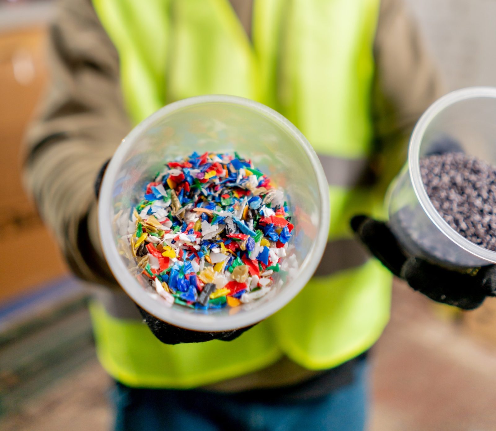 Worker in a high-visibility vest holding out cups of multicolored shredded plastic and dark plastic granules